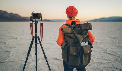 Back view of female tourist with backpack photographing natural landscape of Badwater Basin in California with photo camera on tripod